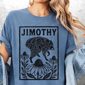 Peut inclure: Un t-shirt bleu avec une impression graphique noire repr&eacute;sentant un raton laveur sautant au-dessus de fleurs. Le texte "JIMOTHY" est imprim&eacute; en caract&egrave;res noirs gras en haut du cadre rectangulaire. Le motif pr&eacute;sente des d&eacute;tails lin&eacute;aires d'un raton laveur.