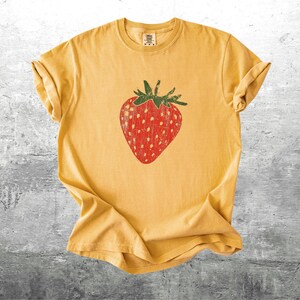 May include: Mustard yellow t-shirt featuring a large, distressed red strawberry graphic with green leaves. The shirt is made of soft material and has rolled-up sleeves. A casual, fruit-themed tee.