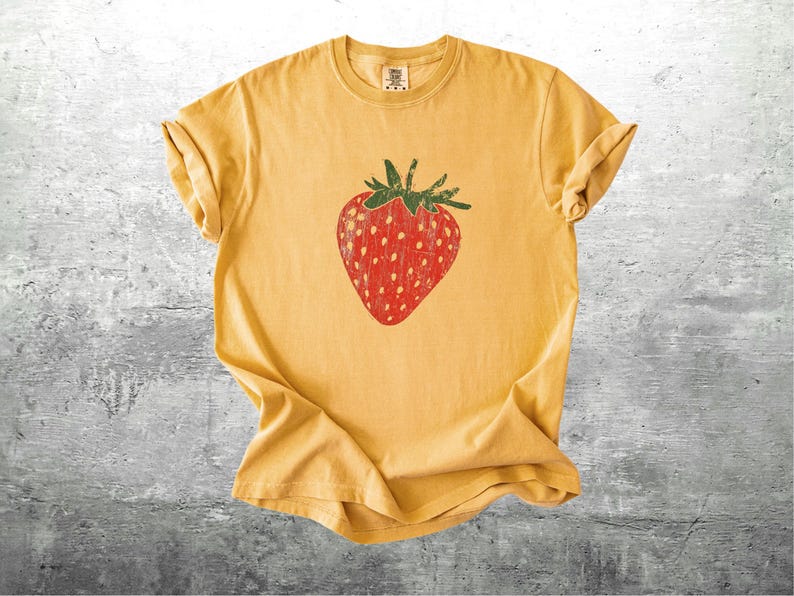 May include: Mustard yellow t-shirt featuring a large, distressed red strawberry graphic with green leaves. The shirt is made of soft material and has rolled-up sleeves. A casual, fruit-themed tee.