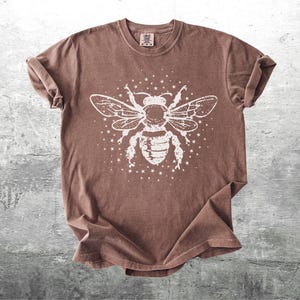 May include: A brown t-shirt featuring a large white graphic of a bee with outstretched wings and surrounding stars. The shirt has a crew neck and short sleeves, with a slightly faded, vintage look.