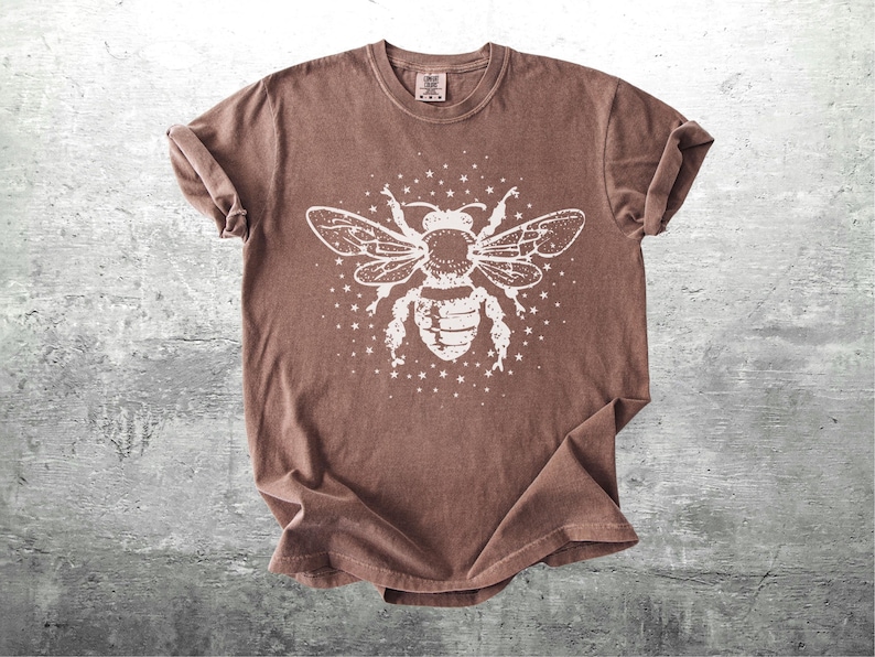 May include: A brown t-shirt featuring a large white graphic of a bee with outstretched wings and surrounding stars. The shirt has a crew neck and short sleeves, with a slightly faded, vintage look.