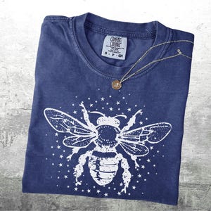 May include: A navy blue t-shirt with a white bee graphic and star accents. A silver necklace with a small pendant rests on the shirt. The shirt is folded and has a Comfort Colors tag.