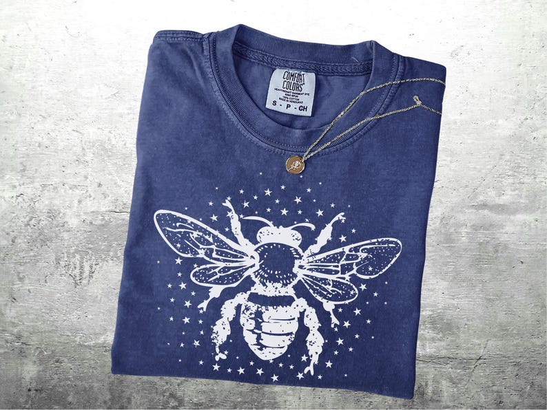 May include: A navy blue t-shirt with a white bee graphic and star accents. A silver necklace with a small pendant rests on the shirt. The shirt is folded and has a Comfort Colors tag.