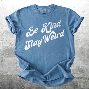 May include: A blue t-shirt with the words "Be Kind Stay Weird" in white, distressed lettering. The shirt is folded, showing the rolled-up sleeves. The background is a gray, textured surface.