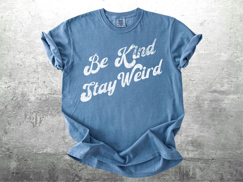 May include: A blue t-shirt with the words "Be Kind Stay Weird" in white, distressed lettering. The shirt is folded, showing the rolled-up sleeves. The background is a gray, textured surface.