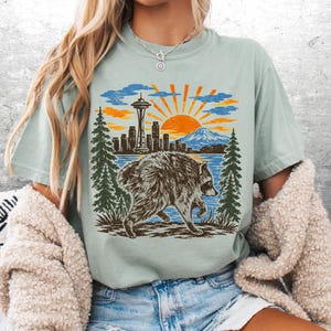 May include: A light sage green t-shirt featuring a graphic print of a raccoon walking in front of a forest, a city skyline with the Space Needle, a mountain, and a bright orange sun. The design uses shades of brown, orange, blue, and green.
