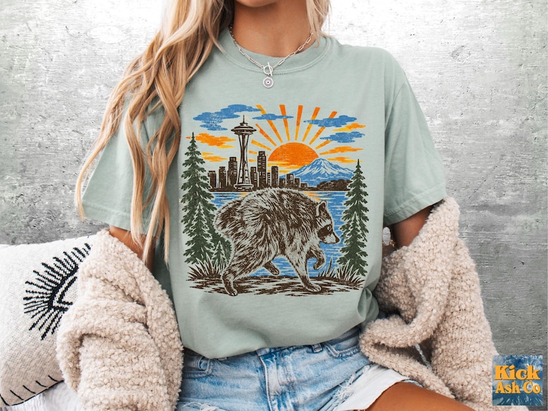 May include: A light sage green t-shirt featuring a graphic print of a raccoon walking in front of a forest, a city skyline with the Space Needle, a mountain, and a bright orange sun. The design uses shades of brown, orange, blue, and green.