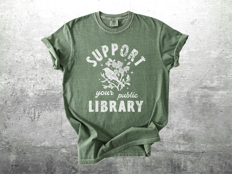 May include: Green t-shirt with the text "SUPPORT your public LIBRARY" in white, along with a bird and floral graphic. The shirt has rolled-up sleeves and a crew neck.