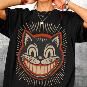 Op de afbeelding: Een zwart t-shirt met korte mouwen en een grote, gecentreerde afbeelding van een gestileerd kattengezicht met een brede, tandige grijns. De kattenillustratie is uitgevoerd in cr&egrave;me- en roestoranje tinten, omringd door uitstralende lijnen.