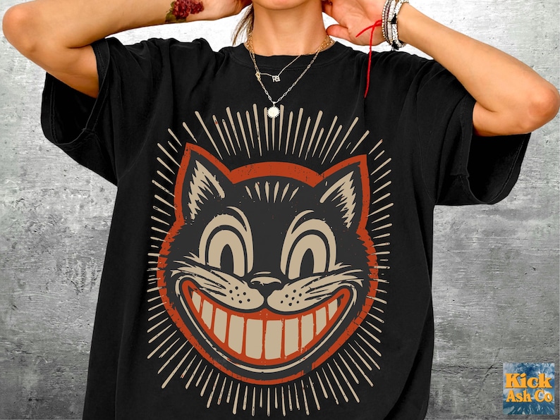 Op de afbeelding: Een zwart t-shirt met korte mouwen en een grote, gecentreerde afbeelding van een gestileerd kattengezicht met een brede, tandige grijns. De kattenillustratie is uitgevoerd in cr&egrave;me- en roestoranje tinten, omringd door uitstralende lijnen.