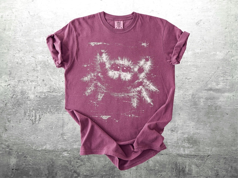 Op de afbeelding: Een bordeauxrood t-shirt met een witte afbeelding van een springspin. De mouwen zijn opgerold. Een klein label is zichtbaar linksboven. De spinnenafbeelding is een centraal ontwerpelement op de voorkant.