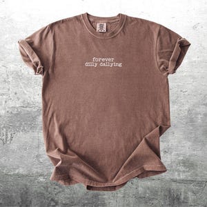 May include: A brown t-shirt with the text "forever dilly dallying" printed in white on the front.