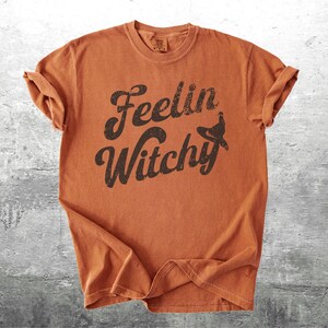 K&ouml;nnte beinhalten: Ein orangefarbenes T-Shirt mit hochgekrempelten &Auml;rmeln. Das Shirt zeigt den Schriftzug "Feelin Witchy" in einer verblassten, dunkelbraunen Schriftart mit einer Hexenhut-Grafik. Das Shirt besteht aus weichem Material und hat einen Vintage-Look.
