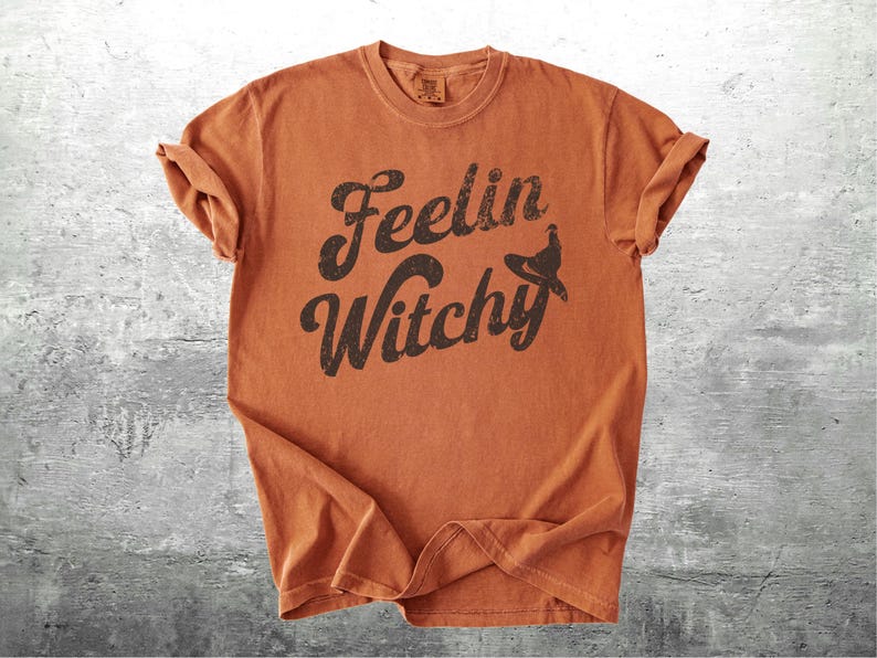 K&ouml;nnte beinhalten: Ein orangefarbenes T-Shirt mit hochgekrempelten &Auml;rmeln. Das Shirt zeigt den Schriftzug "Feelin Witchy" in einer verblassten, dunkelbraunen Schriftart mit einer Hexenhut-Grafik. Das Shirt besteht aus weichem Material und hat einen Vintage-Look.
