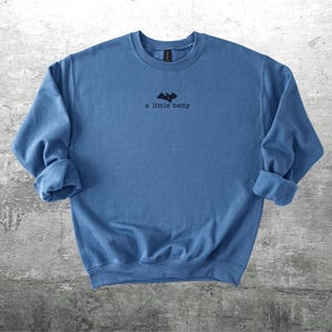 May include: A blue crew neck sweatshirt with rolled-up sleeves. The shirt features a small black bat graphic and the text "a little batty" printed in black. The sweatshirt is displayed on a gray, textured surface.