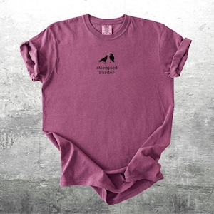 May include: A maroon t-shirt with rolled-up sleeves. The shirt features a small graphic of two black birds and the text "attempted murder" in a simple font. The shirt is displayed on a gray concrete surface.