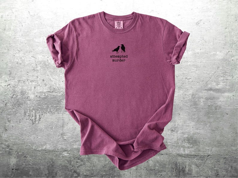 May include: A maroon t-shirt with rolled-up sleeves. The shirt features a small graphic of two black birds and the text "attempted murder" in a simple font. The shirt is displayed on a gray concrete surface.
