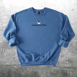 May include: A blue crewneck sweatshirt with the text "cats & coffee" in black. The design includes a cat in a coffee cup. The sleeves are rolled up. The sweatshirt is made of a soft material.