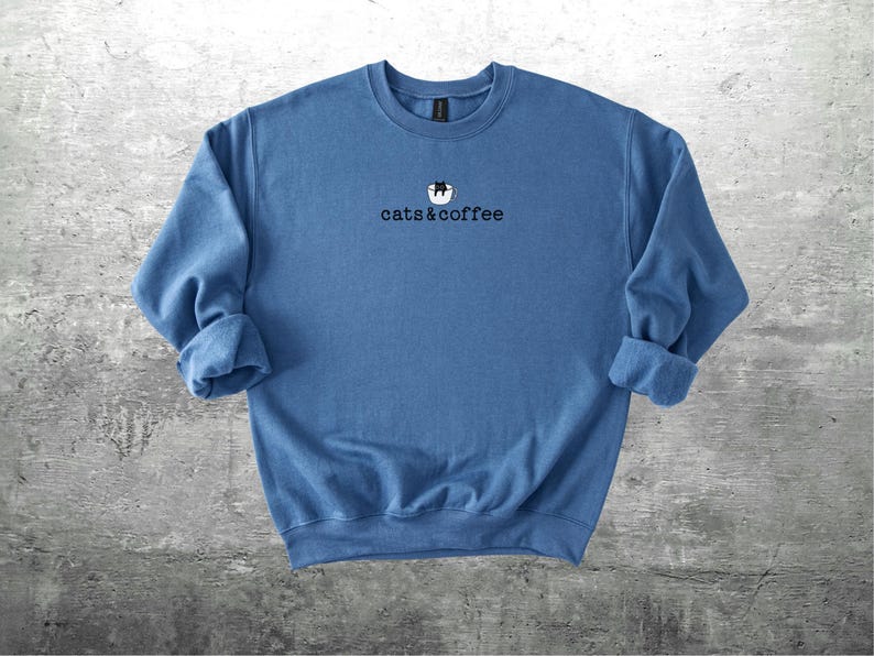 May include: A blue crewneck sweatshirt with the text "cats & coffee" in black. The design includes a cat in a coffee cup. The sleeves are rolled up. The sweatshirt is made of a soft material.