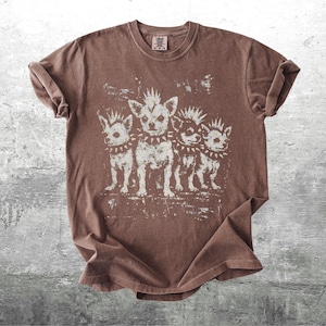 May include: Brown t-shirt featuring a distressed graphic of four dogs with mohawks and studded collars. The dogs are standing in a row, with a vintage, worn look. The shirt has a crew neck and short sleeves.