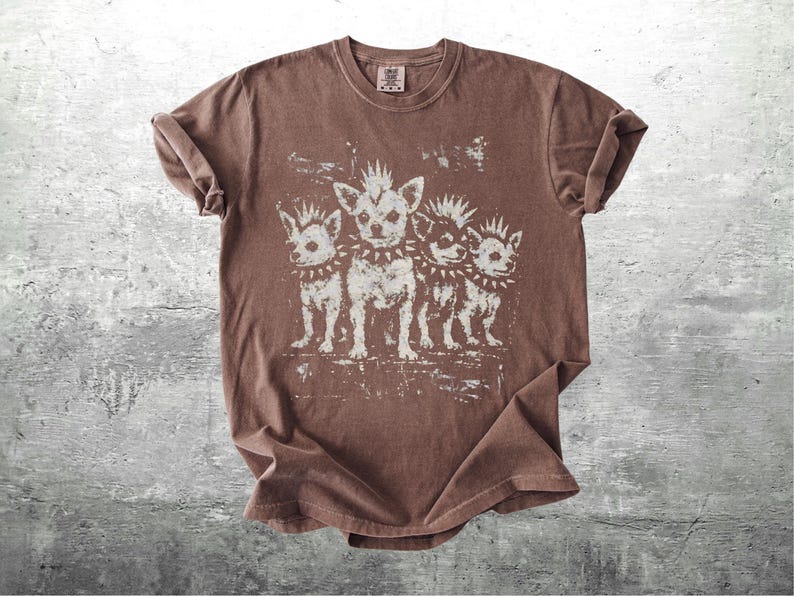 May include: Brown t-shirt featuring a distressed graphic of four dogs with mohawks and studded collars. The dogs are standing in a row, with a vintage, worn look. The shirt has a crew neck and short sleeves.