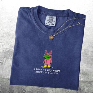 May include: A navy blue Comfort Colors t-shirt featuring a cartoon frog in a pink bunny suit. The text "I have to say weird stuff or I'll die" is printed below the frog. A silver necklace with a pendant is draped across the shirt.