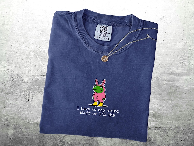 May include: A navy blue Comfort Colors t-shirt featuring a cartoon frog in a pink bunny suit. The text "I have to say weird stuff or I'll die" is printed below the frog. A silver necklace with a pendant is draped across the shirt.