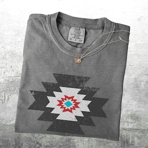 May include: A gray Comfort Colors t-shirt with a geometric design in black, white, red, and turquoise. A silver necklace with a small pendant rests on the shirt. The shirt is folded neatly.