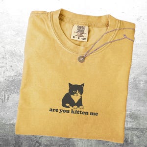 May include: Mustard yellow t-shirt with a black and white cat graphic and the phrase "are you kitten me." A silver necklace with a circular pendant rests on the shirt. The shirt has a Comfort Colors tag.