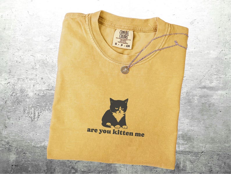 May include: Mustard yellow t-shirt with a black and white cat graphic and the phrase "are you kitten me." A silver necklace with a circular pendant rests on the shirt. The shirt has a Comfort Colors tag.