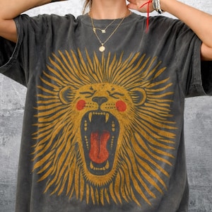 May include: Dark grey t-shirt with a roaring lion design. The lion has a yellow mane, red cheeks, and a red tongue. The shirt has a relaxed fit and short sleeves.