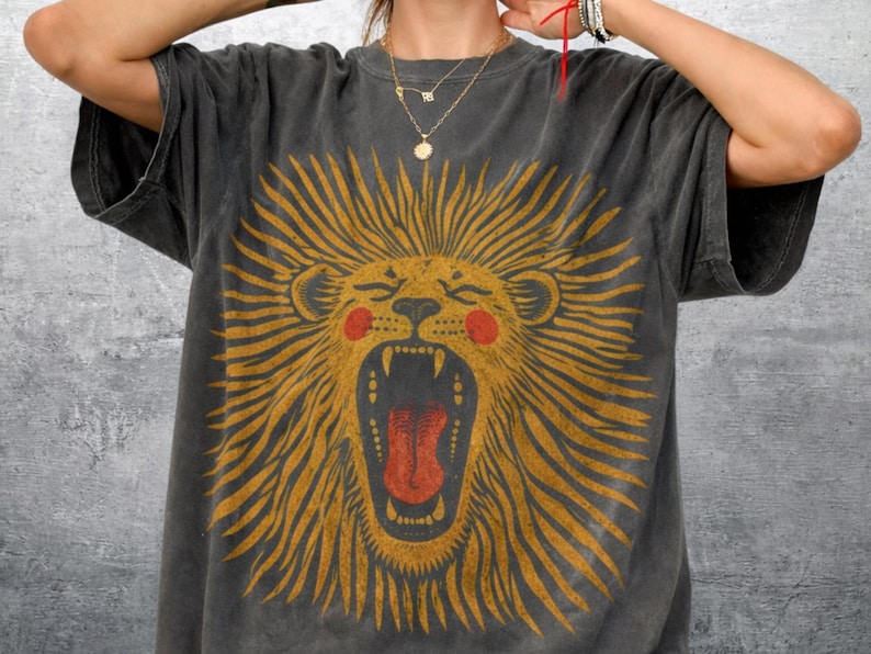 May include: Dark grey t-shirt with a roaring lion design. The lion has a yellow mane, red cheeks, and a red tongue. The shirt has a relaxed fit and short sleeves.