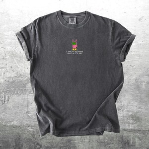 May include: Dark grey t-shirt featuring a frog in a pink bunny suit graphic. The text "I have to say weird stuff or I'll die" is printed below the graphic. The shirt has rolled-up sleeves.