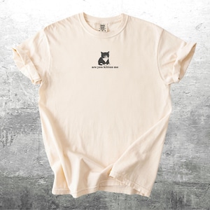 May include: Cream-colored t-shirt with a black graphic of a cat and the phrase "are you kitten me" printed below. The shirt has a crew neck and short sleeves. The fabric is slightly wrinkled, giving it a casual look.
