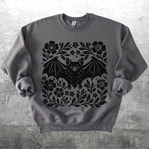 May include: A gray sweatshirt featuring a black bat design surrounded by flowers and leaves. The bat has its wings spread, and the floral design is symmetrical. The sweatshirt is made of a soft material.