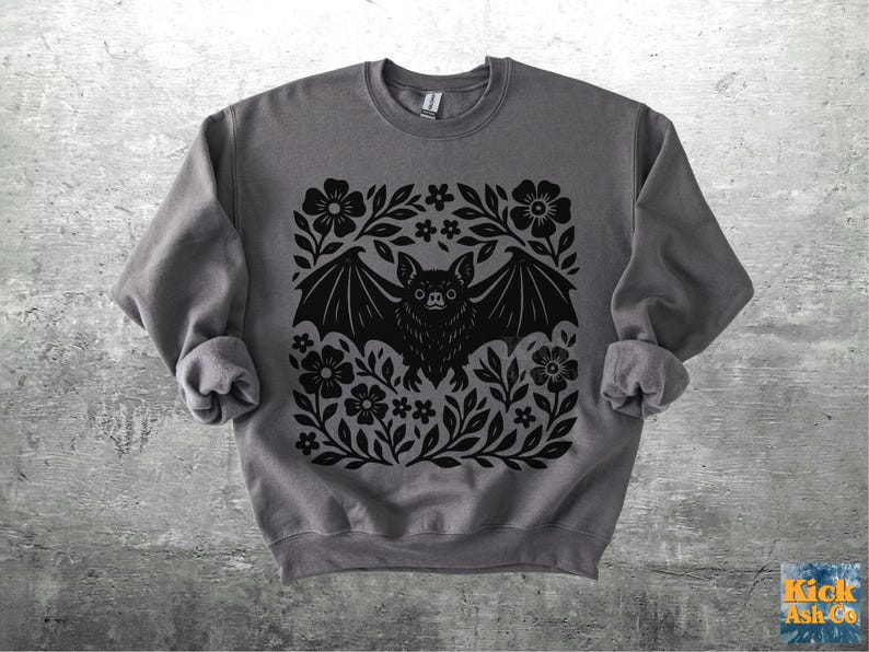 May include: A gray sweatshirt featuring a black bat design surrounded by flowers and leaves. The bat has its wings spread, and the floral design is symmetrical. The sweatshirt is made of a soft material.
