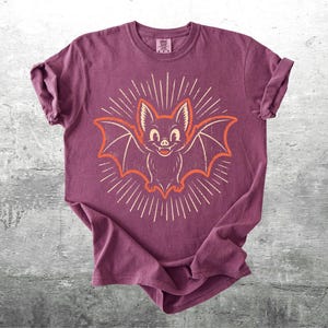 May include: A plum-colored short-sleeve t-shirt featuring a central graphic of a smiling bat with outstretched wings. The bat is outlined in orange and surrounded by a sunburst pattern of thin white lines radiating outward from the center.