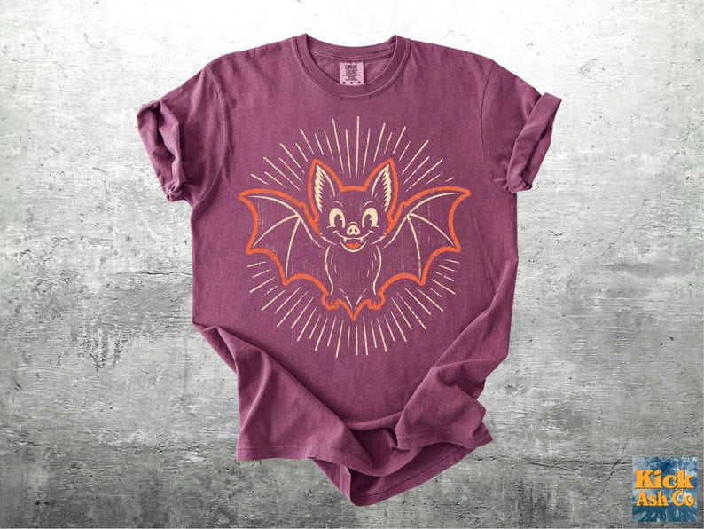 May include: A plum-colored short-sleeve t-shirt featuring a central graphic of a smiling bat with outstretched wings. The bat is outlined in orange and surrounded by a sunburst pattern of thin white lines radiating outward from the center.