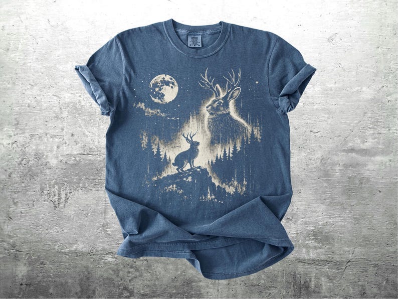 May include: A blue t-shirt featuring a graphic of a jackalope with antlers, a full moon, and a forest scene. The design is in shades of beige and white, creating a vintage aesthetic. The shirt is made of soft material.
