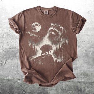 K&ouml;nnte beinhalten: Ein braunes Kurzarm-T-Shirt mit einem wei&szlig;en Grafikdruck eines Waschb&auml;rengesichts, eines Vollmonds und eines B&auml;ren, der auf einer felsigen Klippe inmitten eines Waldes l&auml;uft. Das T-Shirt hat einen Rundhalsausschnitt und ein Etikett am Innenkragen.