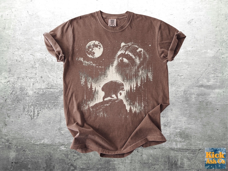 K&ouml;nnte beinhalten: Ein braunes Kurzarm-T-Shirt mit einem wei&szlig;en Grafikdruck eines Waschb&auml;rengesichts, eines Vollmonds und eines B&auml;ren, der auf einer felsigen Klippe inmitten eines Waldes l&auml;uft. Das T-Shirt hat einen Rundhalsausschnitt und ein Etikett am Innenkragen.