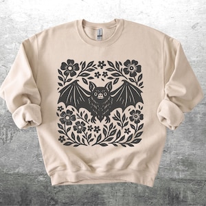 May include: Beige sweatshirt featuring a black bat design surrounded by flowers and leaves. The bat has detailed wings and a focused expression. The design is centered on the front of the long-sleeved crew neck sweatshirt.