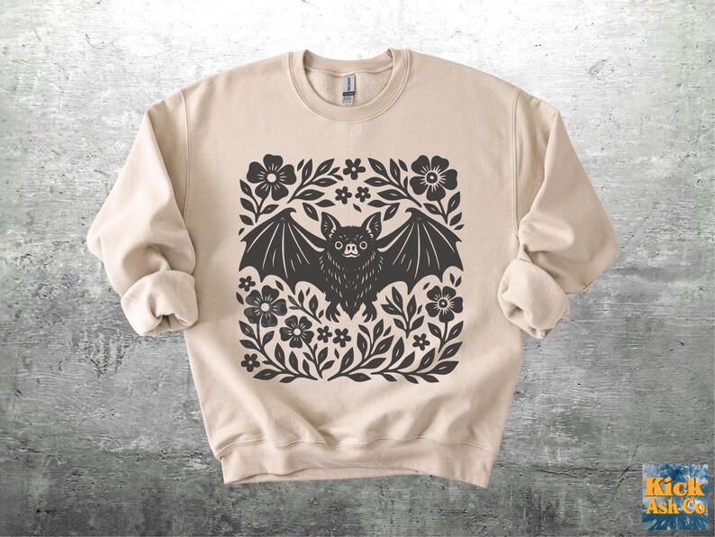 May include: Beige sweatshirt featuring a black bat design surrounded by flowers and leaves. The bat has detailed wings and a focused expression. The design is centered on the front of the long-sleeved crew neck sweatshirt.