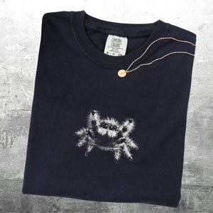 May include: A navy blue t-shirt with a white spider graphic. A gold necklace with a small pendant rests on the shirt. The shirt has a Comfort Colors tag.