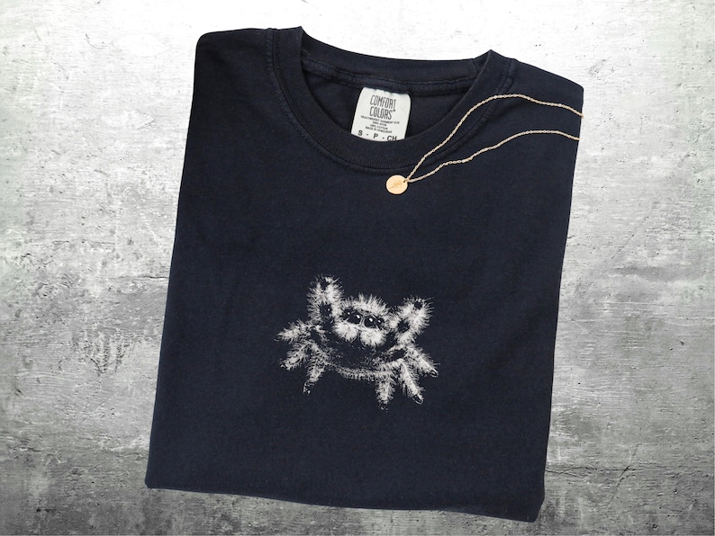 May include: A navy blue t-shirt with a white spider graphic. A gold necklace with a small pendant rests on the shirt. The shirt has a Comfort Colors tag.