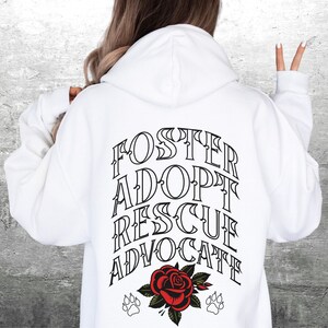 Pu&ograve; includere: Felpa con cappuccio bianca con il testo "FOSTER ADOPT RESCUE ADVOCATE" stampato sul retro. Un rosa rossa con foglie verdi e due impronte di zampa si trovano sotto il testo.