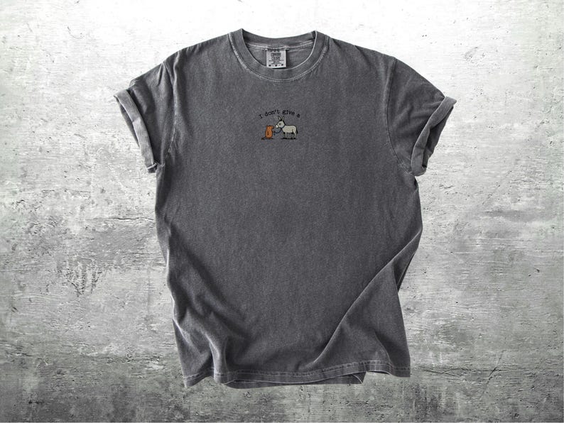May include: Charcoal grey t-shirt with rolled sleeves. The shirt has an embroidered design of a donkey and a barrel, with the text "I don't give a". The fabric has a soft, textured appearance.