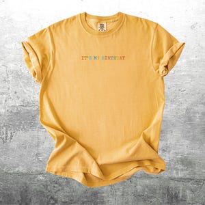 Puede incluir: Una camiseta amarilla con el texto "It's My Birthday" en colores arco&iacute;ris.