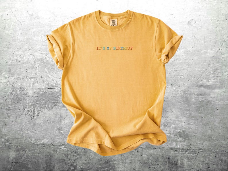Puede incluir: Una camiseta amarilla con el texto "It's My Birthday" en colores arco&iacute;ris.