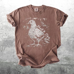 May include: Brown t-shirt with a distressed white graphic of a pigeon and the text "that's Coo". The shirt has rolled-up sleeves and a crew neck. The shirt is made of a soft material.